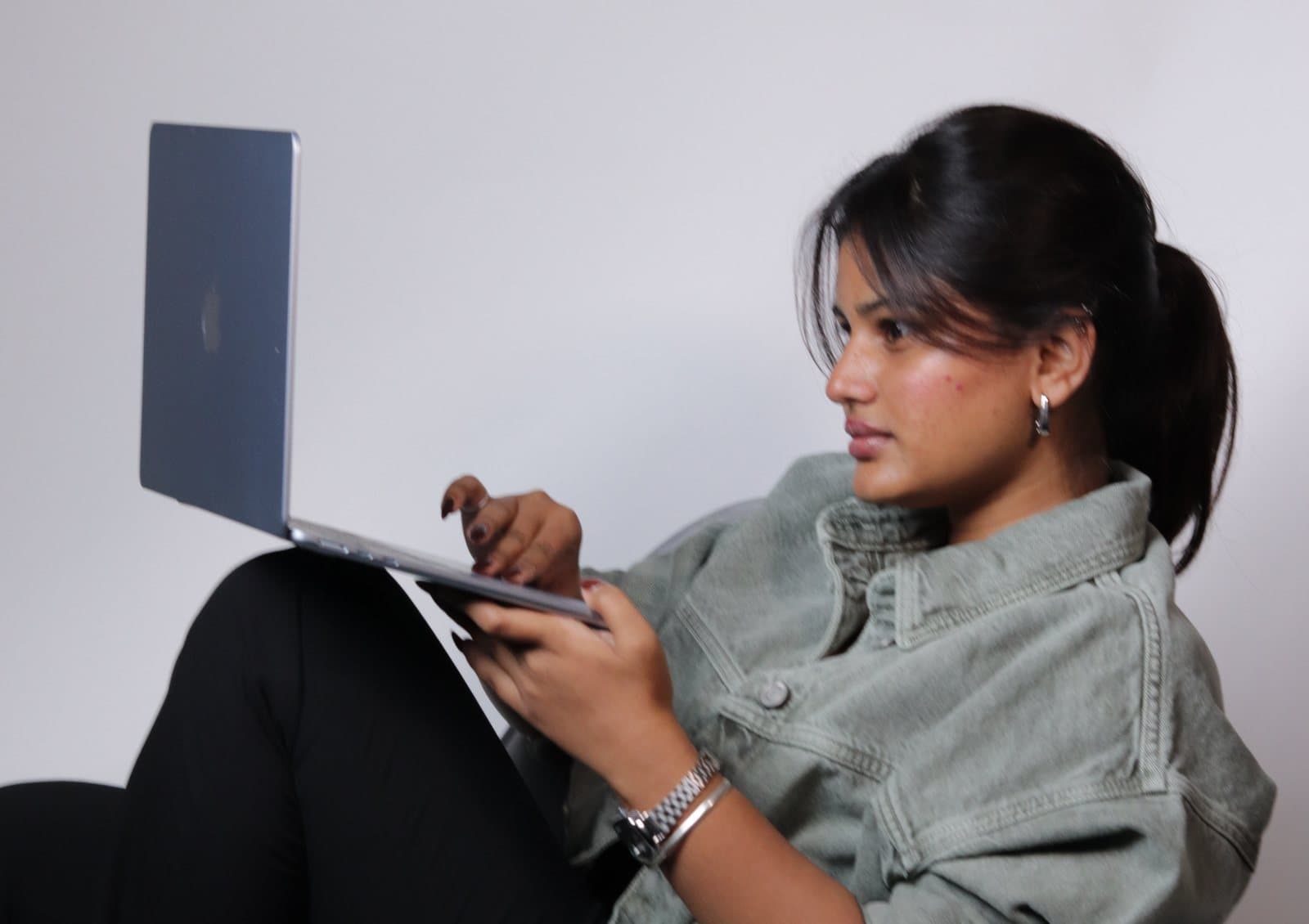 Swapnaja in a sage denim jacket, curled up in an armchair, working on a laptop.