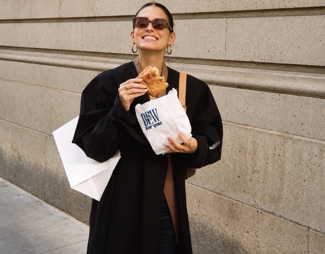 A woman on the street holding a croissant and a Brew for You paper bag