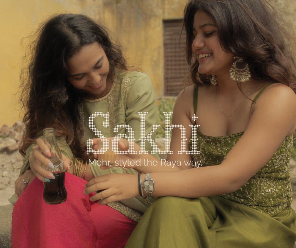 Two women laughing in festive wear, one holding a bottle of cola, under the title “Sakhi: Mehr, styled the Raya way.”