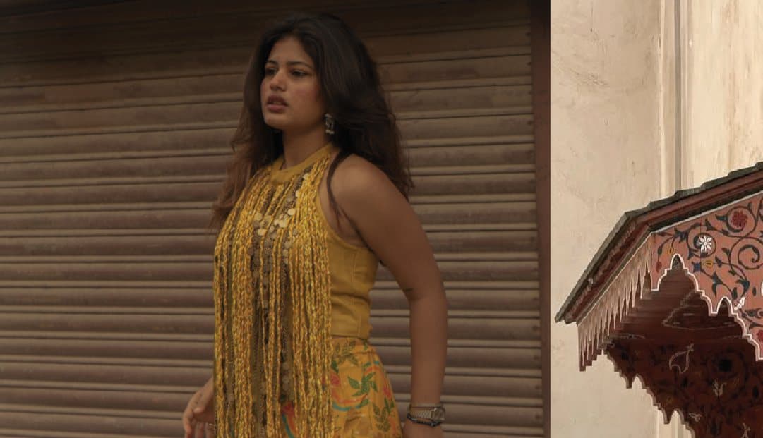 A woman in a mustard fringed top before a rolled-down shutter, beside a carved wooden eave