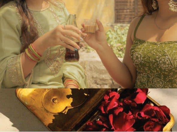 Two women toasting with a bottle of cola and a glass of chai, above a brass tray of red rose petals