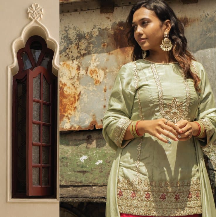 A woman in a sage embroidered kurta beside an arched window in a weathered wall