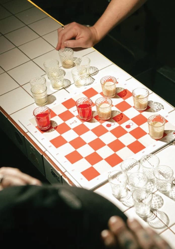 Hands over a drinking-game chessboard of shot glasses