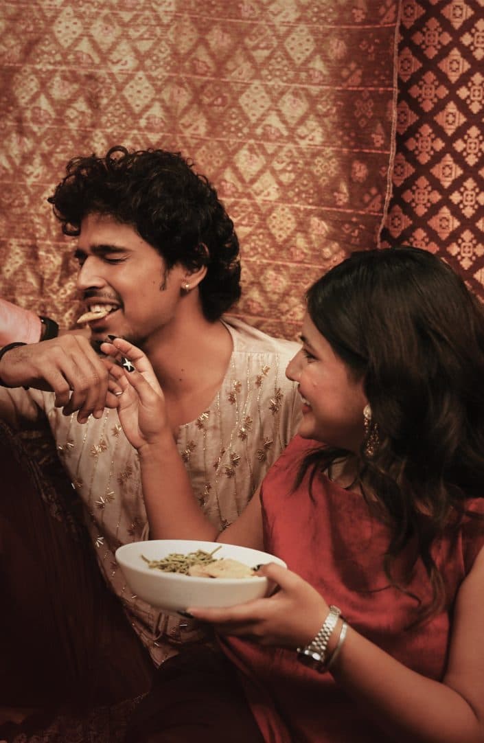 A woman feeding a laughing man from a bowl, before a patterned hanging