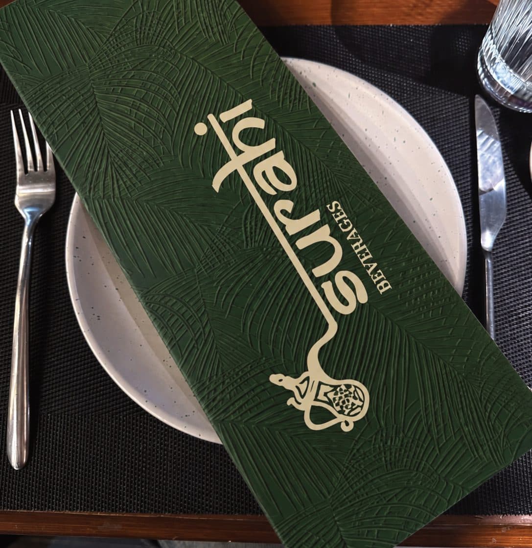 The Surahi beverage menu, an embossed green cover with the logo in cream, laid on a dinner plate