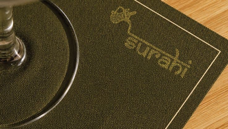 An olive napkin printed with the Surahi logo, under the foot of a glass