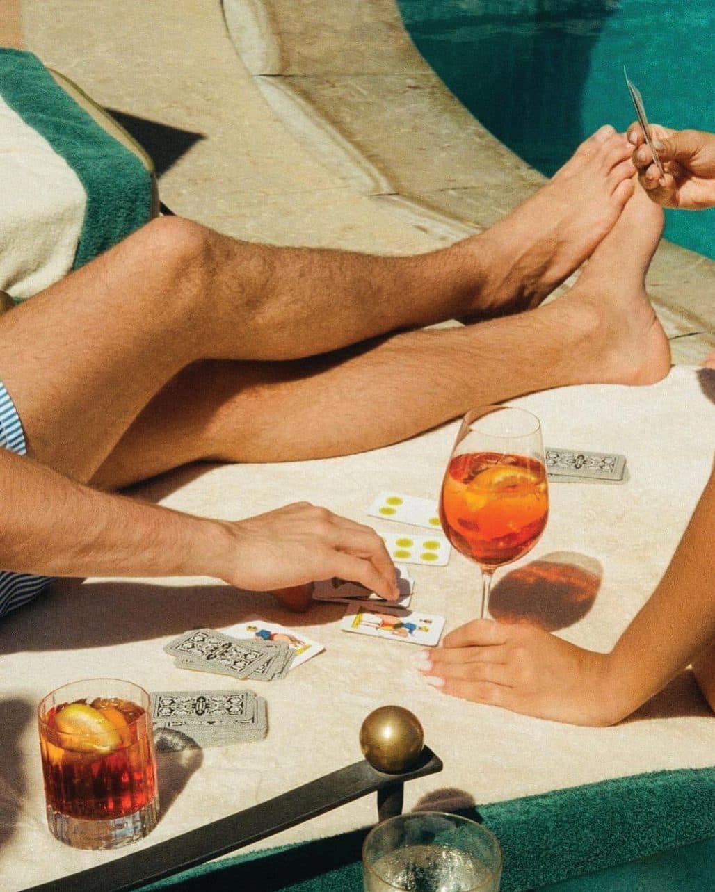 Legs stretched out on a poolside lounger, beside an aperitif and a deck of cards