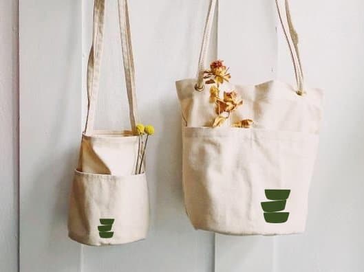 Two canvas bags printed with the logomark, hung on a white wall with dried flowers